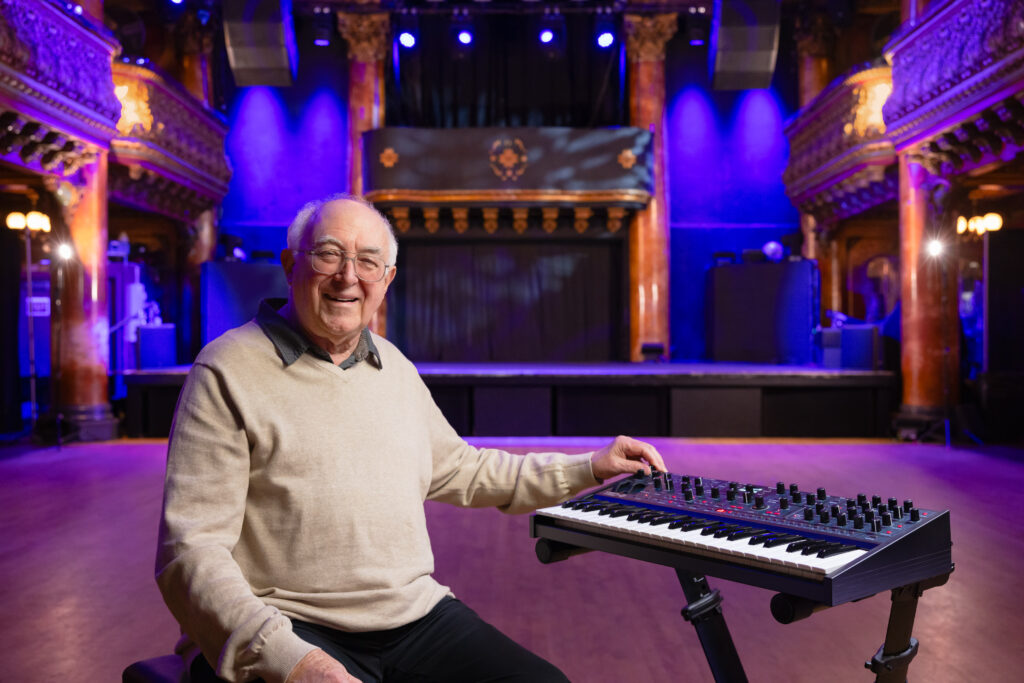 Tom Oberheim is smiling, seated next to a TEO-5 synthesizer at the Great American Music Hall, San Francisco.